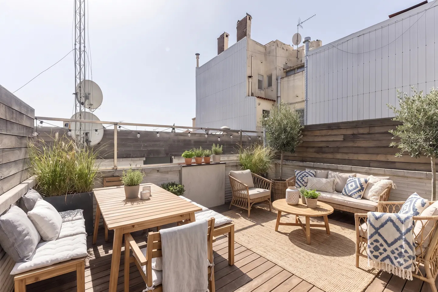 Outdoor rooftop patio with wooden deck, table, chairs, sofa, and plants. A woven rug sits under a round coffee table. Buildings are visible in the background.