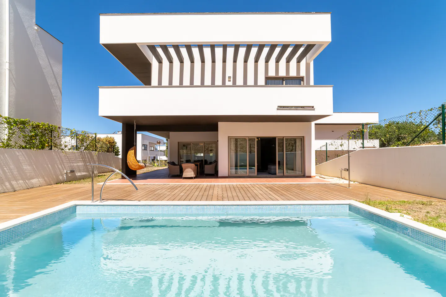 Modern white two-story villa with a wooden deck, swimming pool, and hanging chair under a clear blue sky.