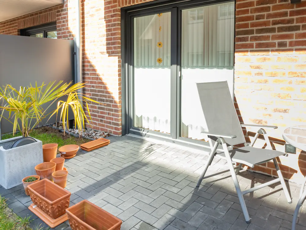 A patio with gray pavers, a gray chair, and terracotta pots. A brick wall and glass doors are in the background.