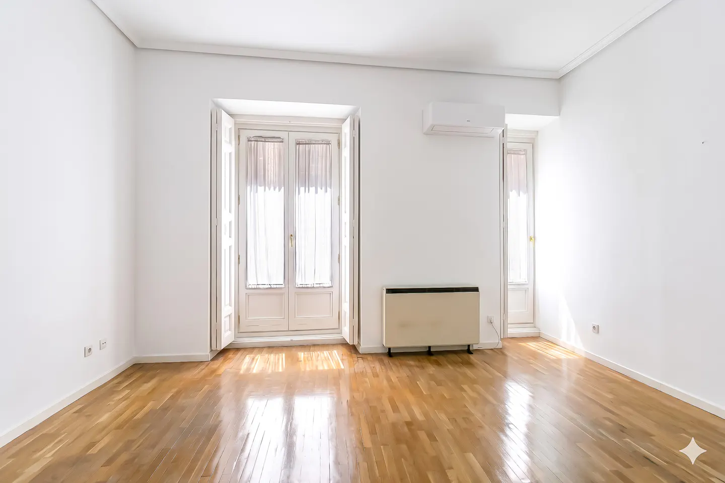 Empty room with white walls, wood floors, and two sets of white doors with sheer curtains. A heater and AC unit are on the wall.
