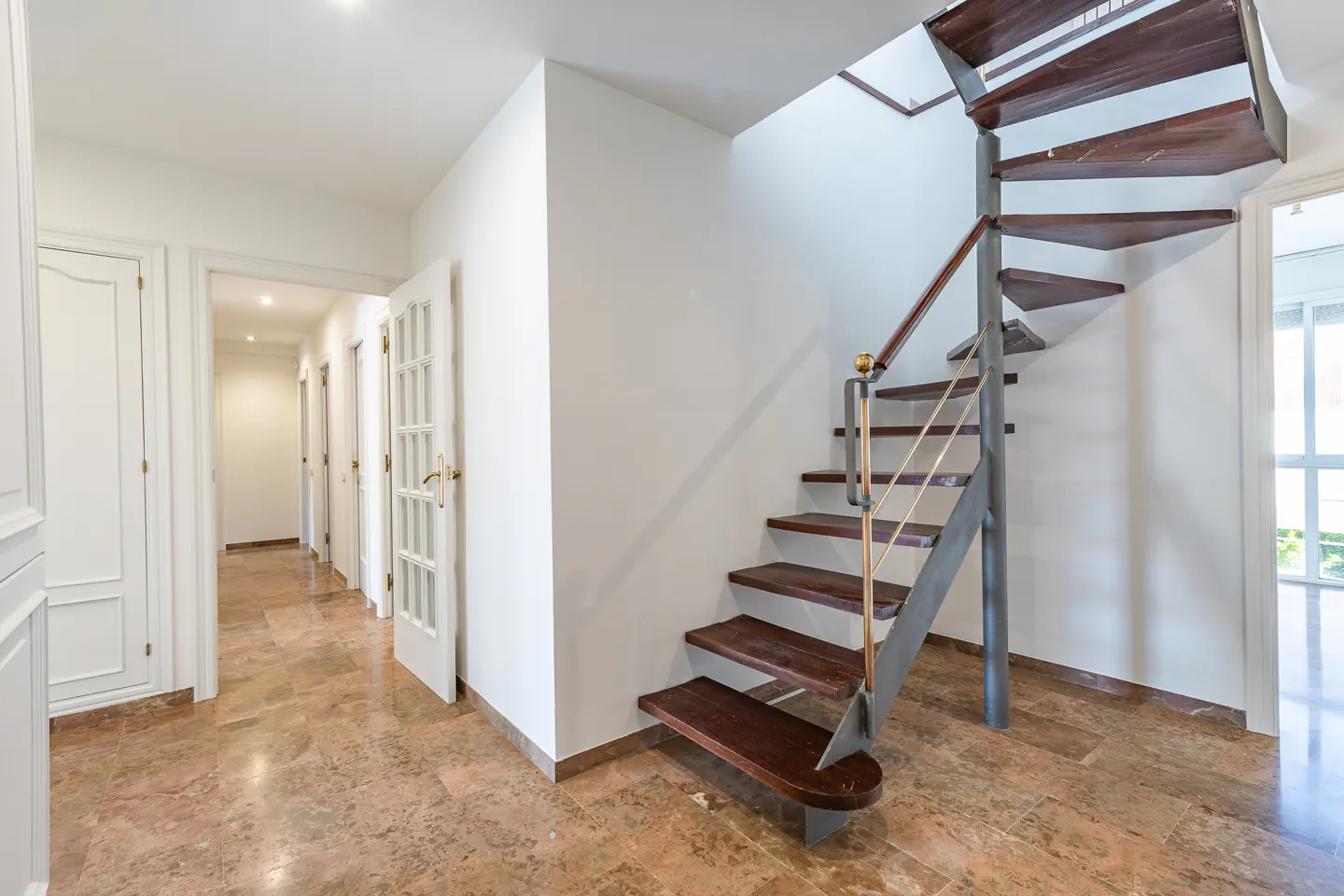 Hallway with a spiral staircase. The stairs are wood with a metal railing. White walls and marble floors.
