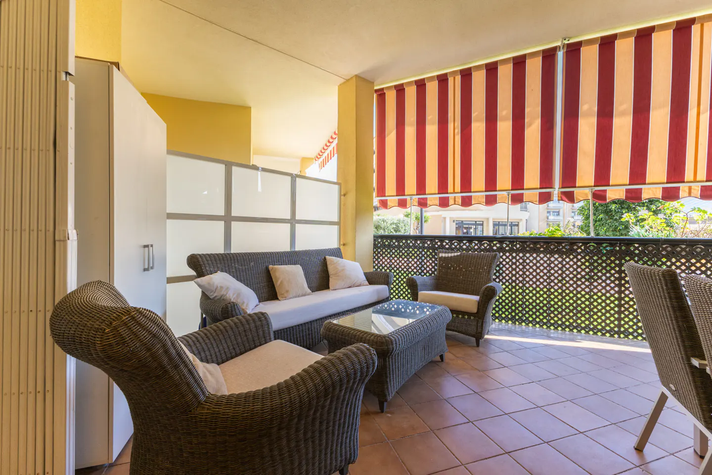 A covered patio with wicker furniture, including a sofa, chairs, and a glass-topped table, under a red and tan striped awning.