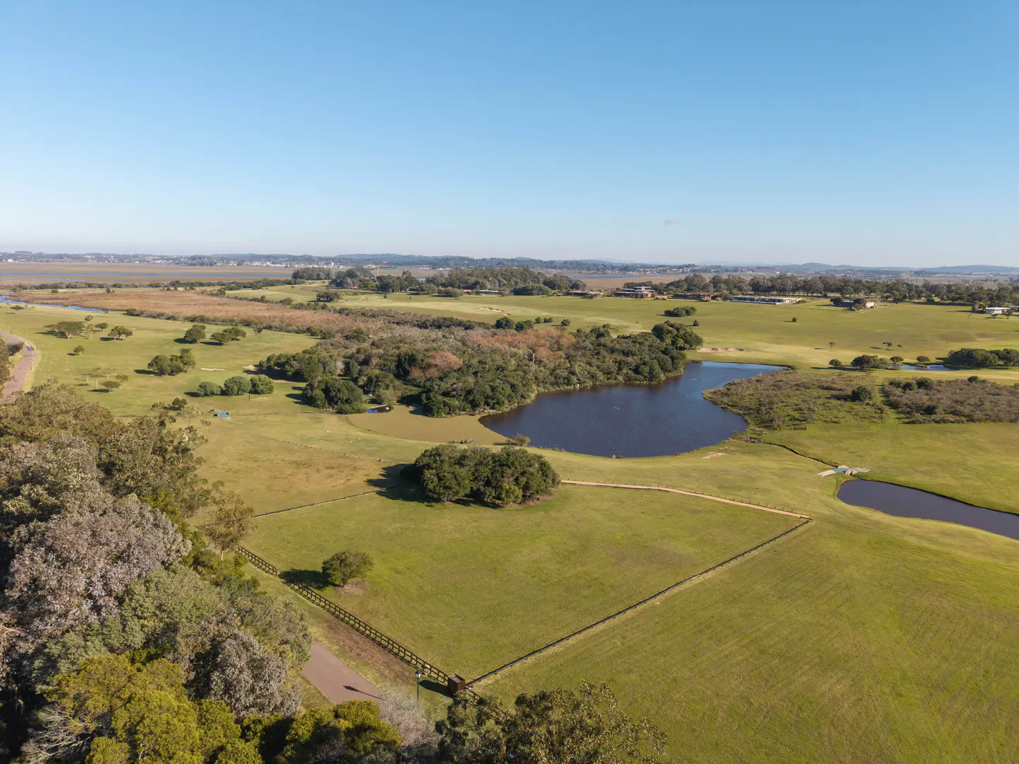 Aerial view of a green field with a dark blue pond, trees, and a wooden fence under a clear blue sky.
