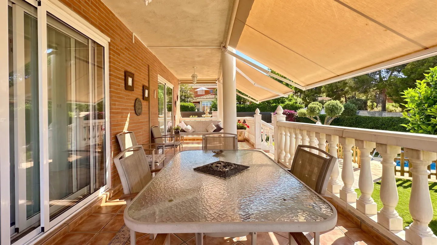 Spacious covered terrace with a glass dining table, brick wall, and white balustrade overlooking a garden.
