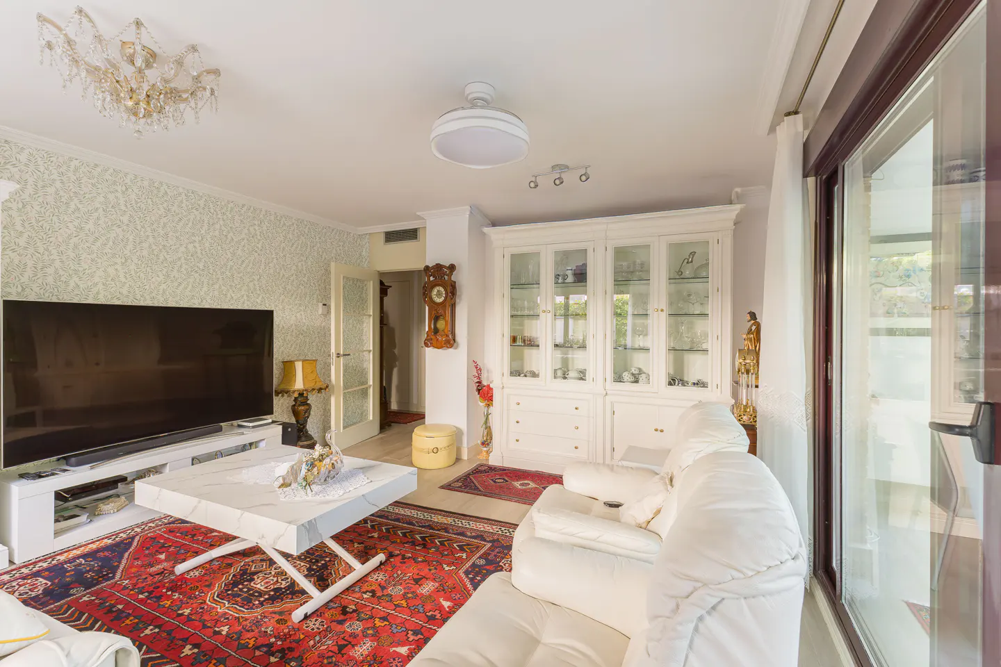 Living room with white sofa, marble table on red rug, TV, and white cabinet. Chandelier and natural light fill the space.