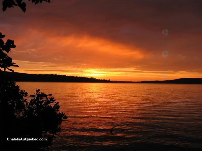 Sunset over a calm lake, reflecting orange and red hues. Silhouetted trees line the horizon and foreground.