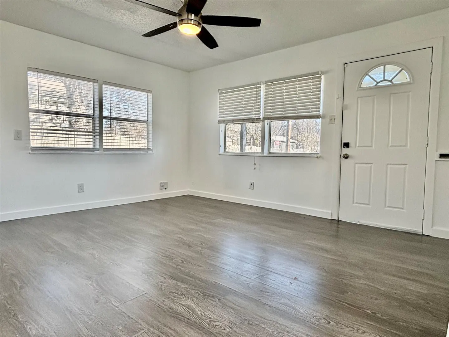 Empty room with gray wood-look floors, white walls, two windows with blinds, and a white door. A ceiling fan is visible.