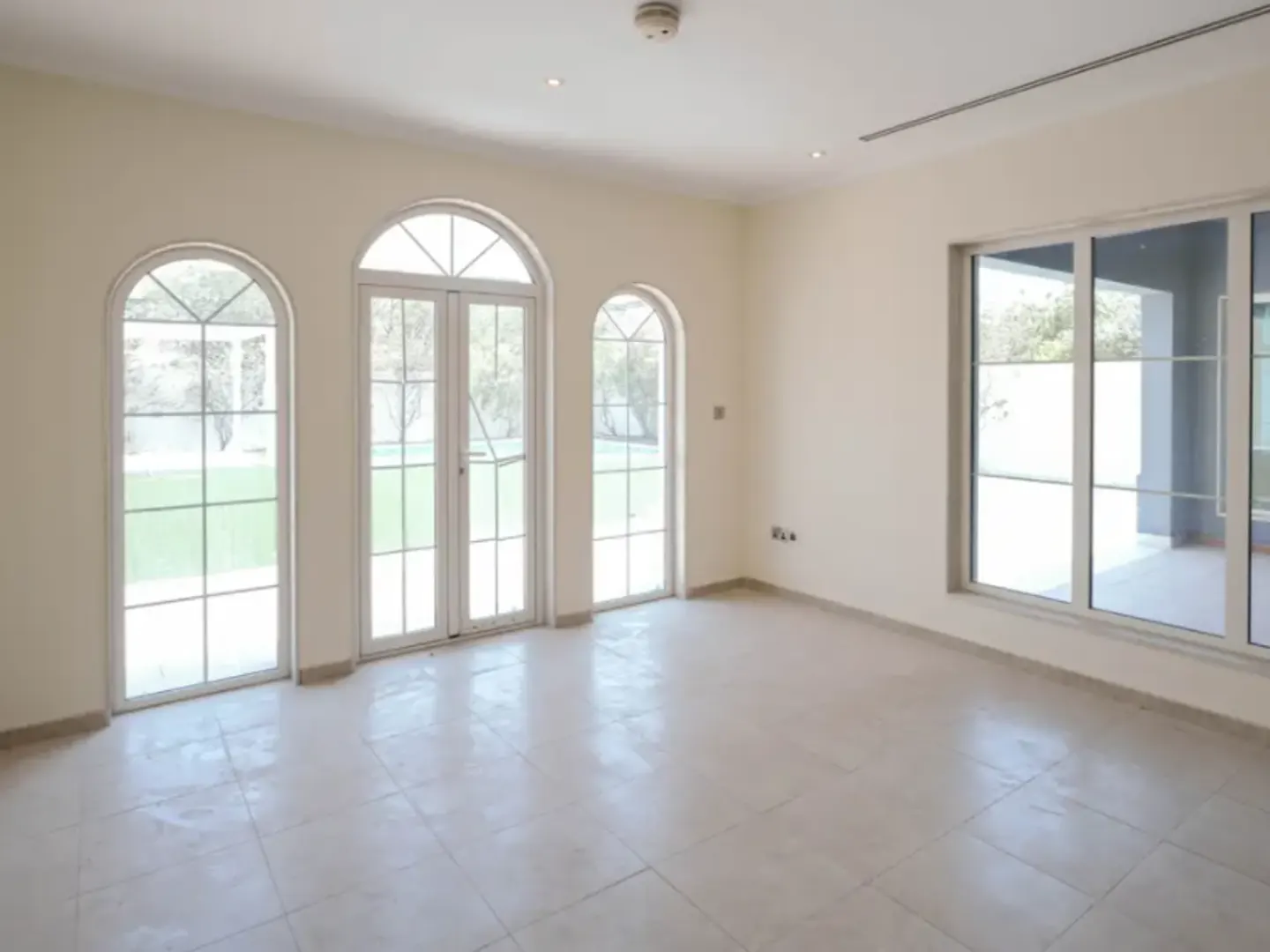 Empty room with cream walls, tiled floor, arched windows and glass doors leading to a green lawn.