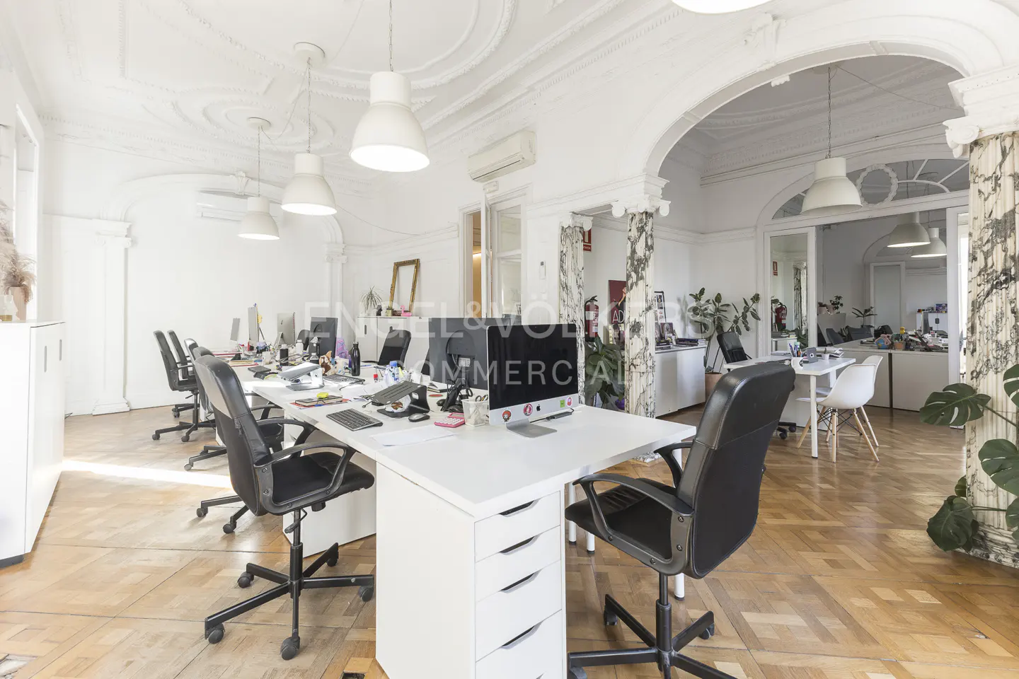 Bright office space with white walls, ornate ceilings, and wood floors. Desks with computers and black chairs are arranged in rows.