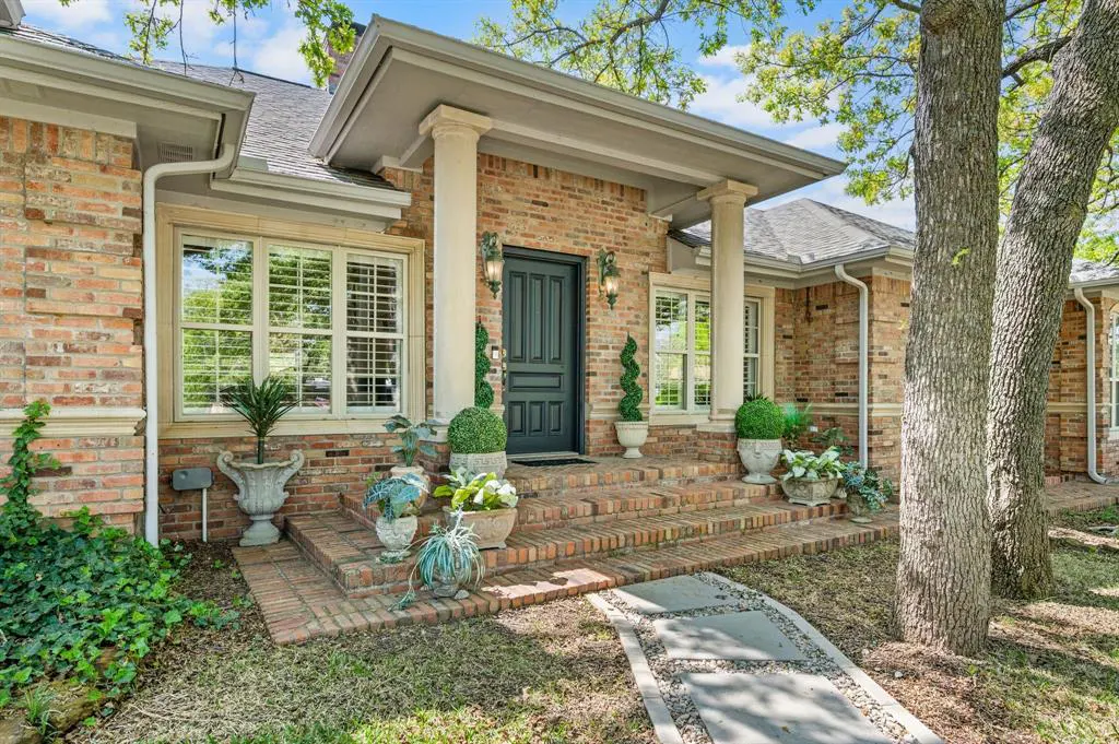 Brick home exterior with a dark green front door, white columns, and potted plants on the steps.