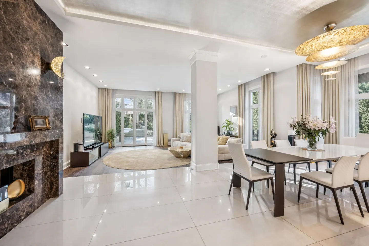 Modern open-plan living and dining area with white tile floors, a dark marble fireplace, and elegant gold pendant lighting.