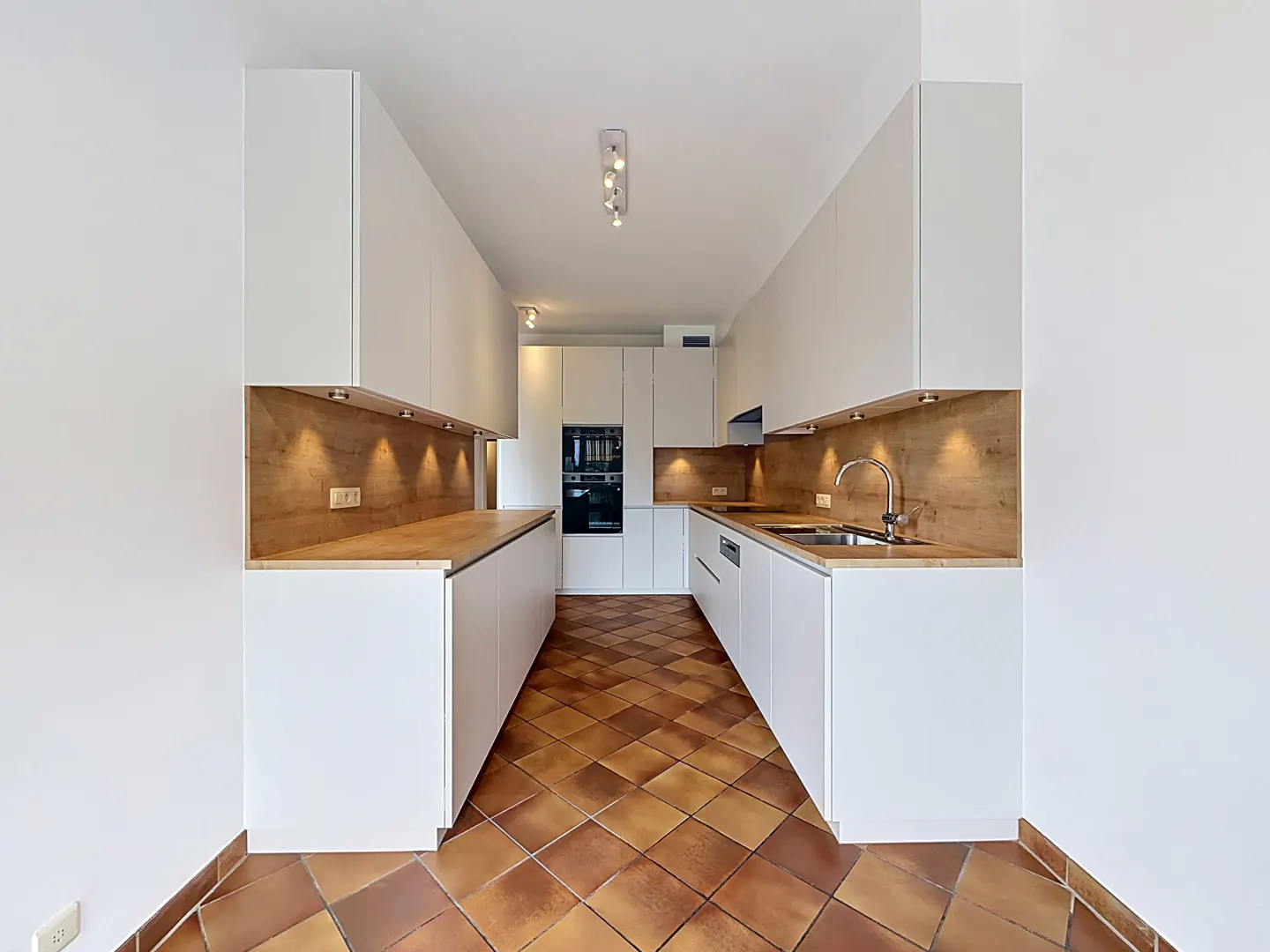 Modern galley kitchen with white cabinets, wood countertops, under-cabinet lighting, and brown diamond-pattern tile floors.