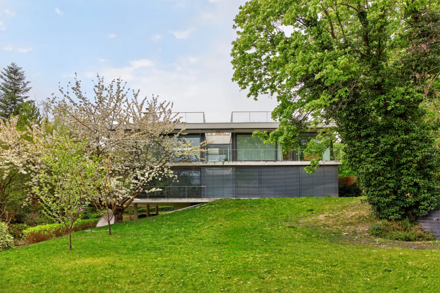 Two-story modern house with large windows and a flat roof, surrounded by green trees and a grassy lawn.