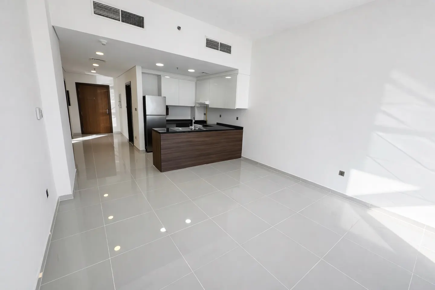 A bright, empty apartment with gray tile floors and a modern kitchen. The kitchen has white cabinets and a dark wood island.