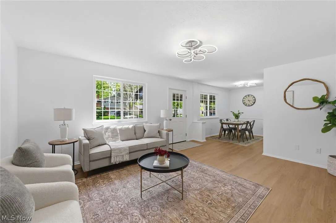 Bright living room with white walls, wood floors, and a patterned rug. A gray sofa and white chairs surround a black coffee table. A dining area is visible in the background.