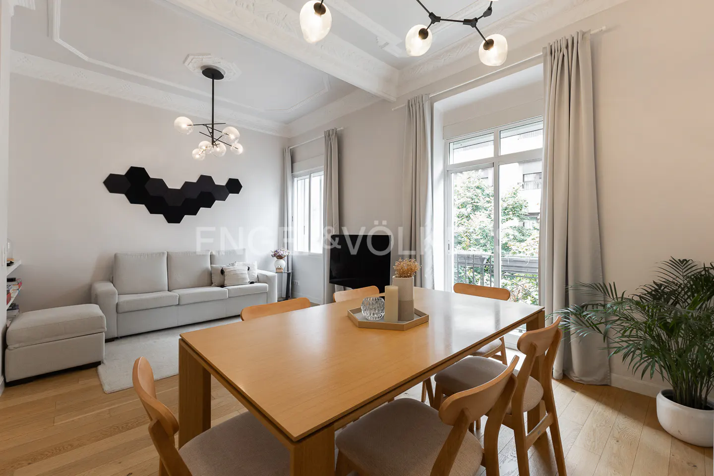 Bright living room with a wooden table, chairs, sofa, and black hexagon wall decor. Large windows with light gray curtains let in natural light.