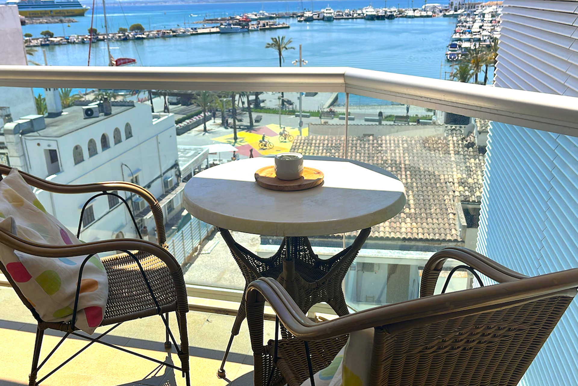 Balcony view with two wicker chairs, a small round table with a coffee cup, overlooking a harbor with boats and buildings.