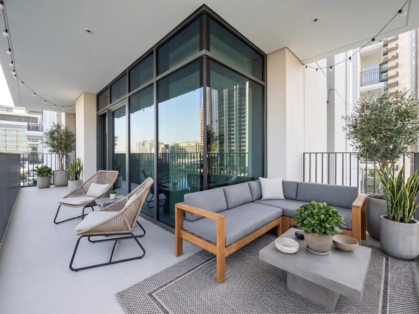 A modern balcony with gray cushioned seating, woven chairs, and potted plants. A gray rug and coffee table complete the outdoor living space.