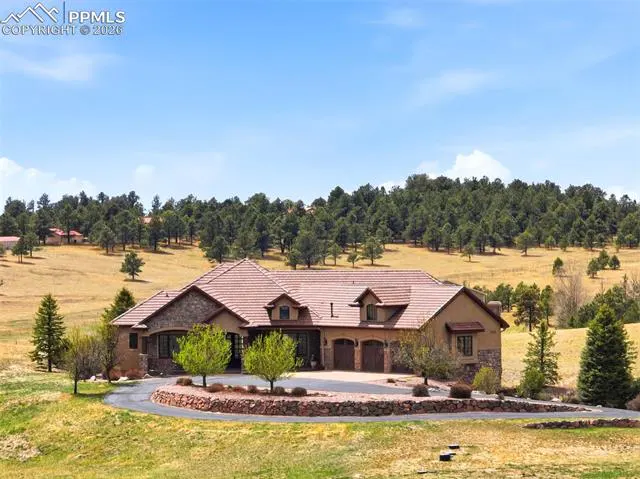 A large, tan house with a red tile roof sits on a green lawn with trees in the background.