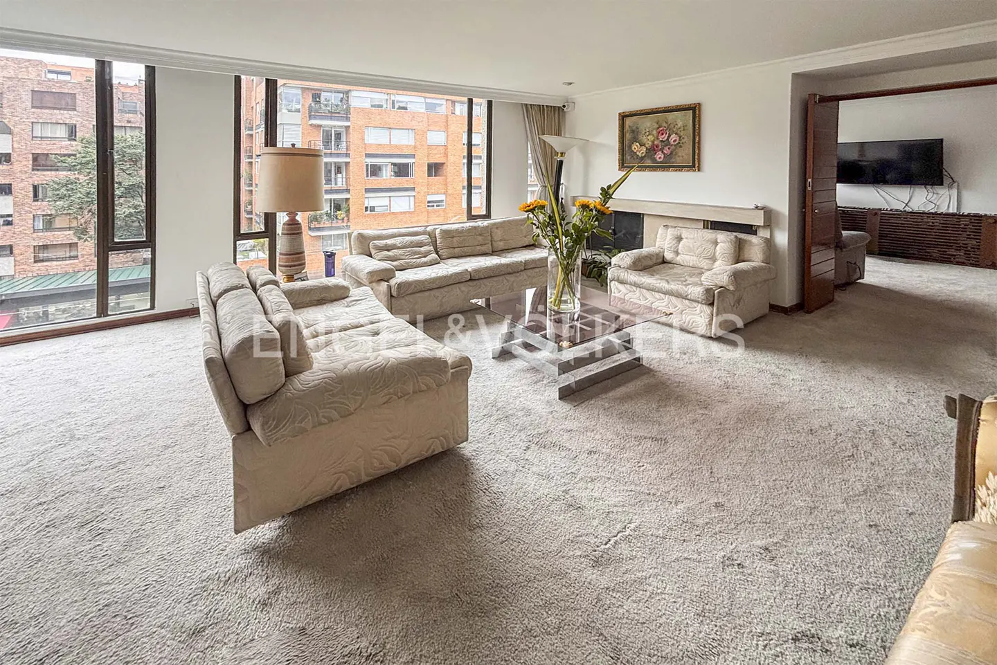 Living room with beige sofas, a glass coffee table, and large windows overlooking a city. A fireplace and floral painting add warmth.