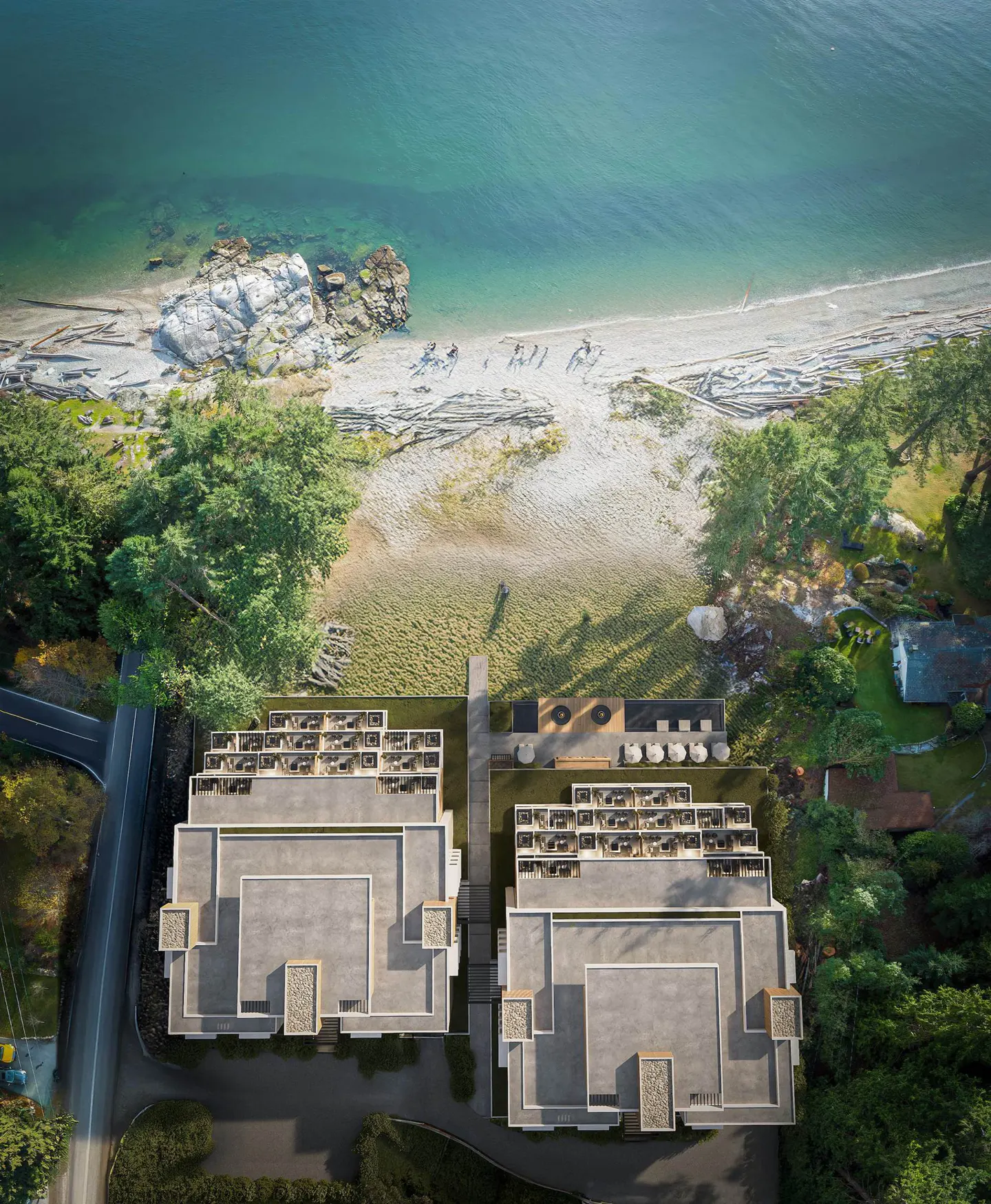 Aerial view of two modern buildings with patios, near a sandy beach and turquoise ocean. Trees surround the buildings.