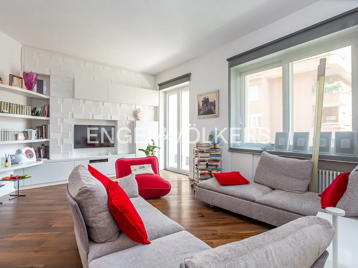 A modern living room with gray sofas, red pillows, and a white wall unit with a TV. Hardwood floors and large windows add to the bright, airy feel.