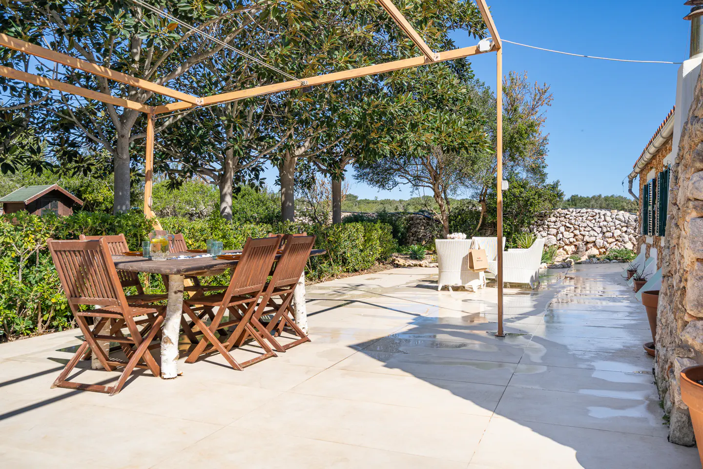 Outdoor patio with a wooden table and chairs under a pergola, with white wicker chairs nearby. Lush greenery and a stone wall in the background.