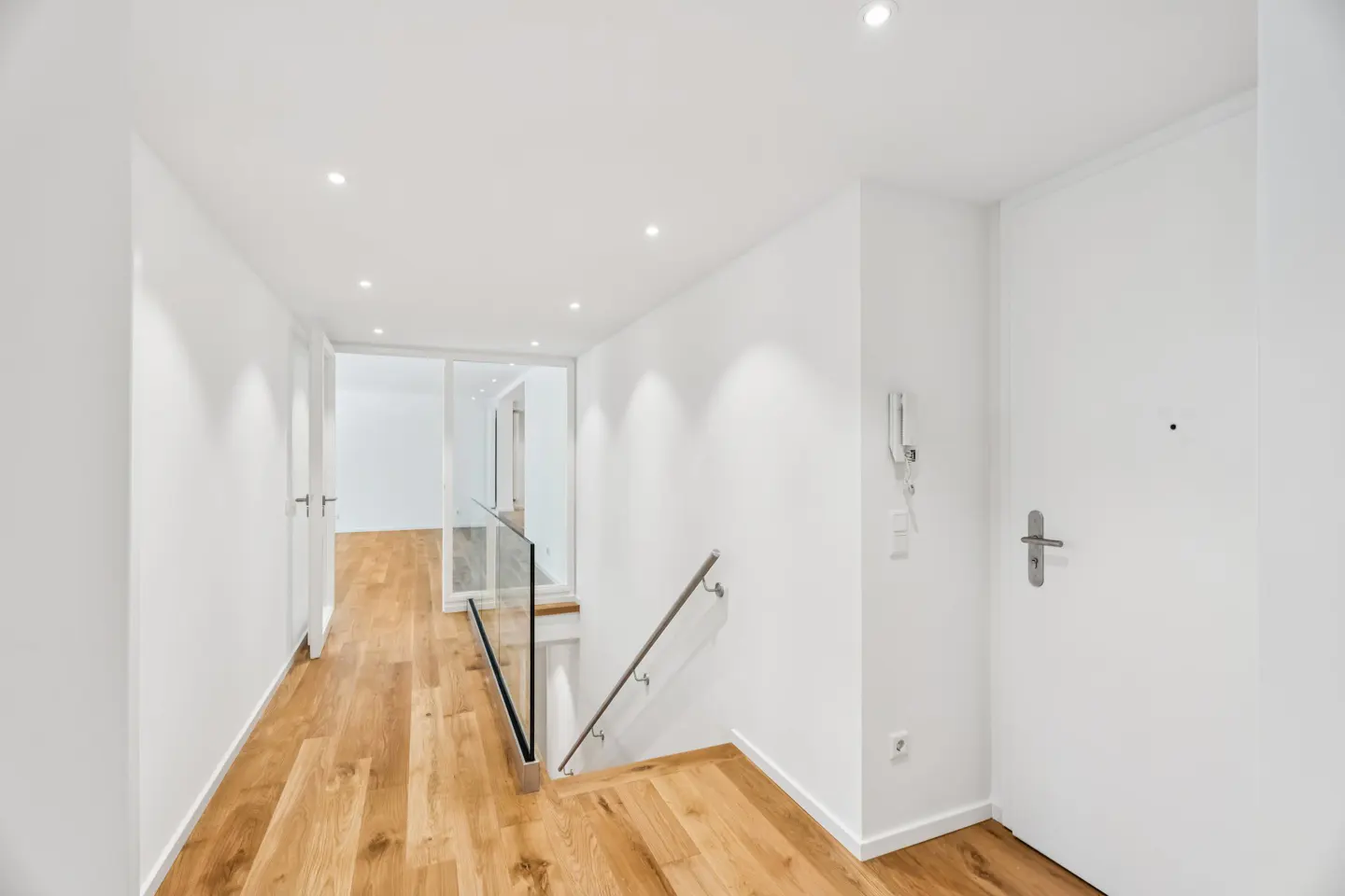 Bright, modern hallway with white walls, wood floors, and recessed lighting. A glass-paneled staircase leads down. A white door is on the right.