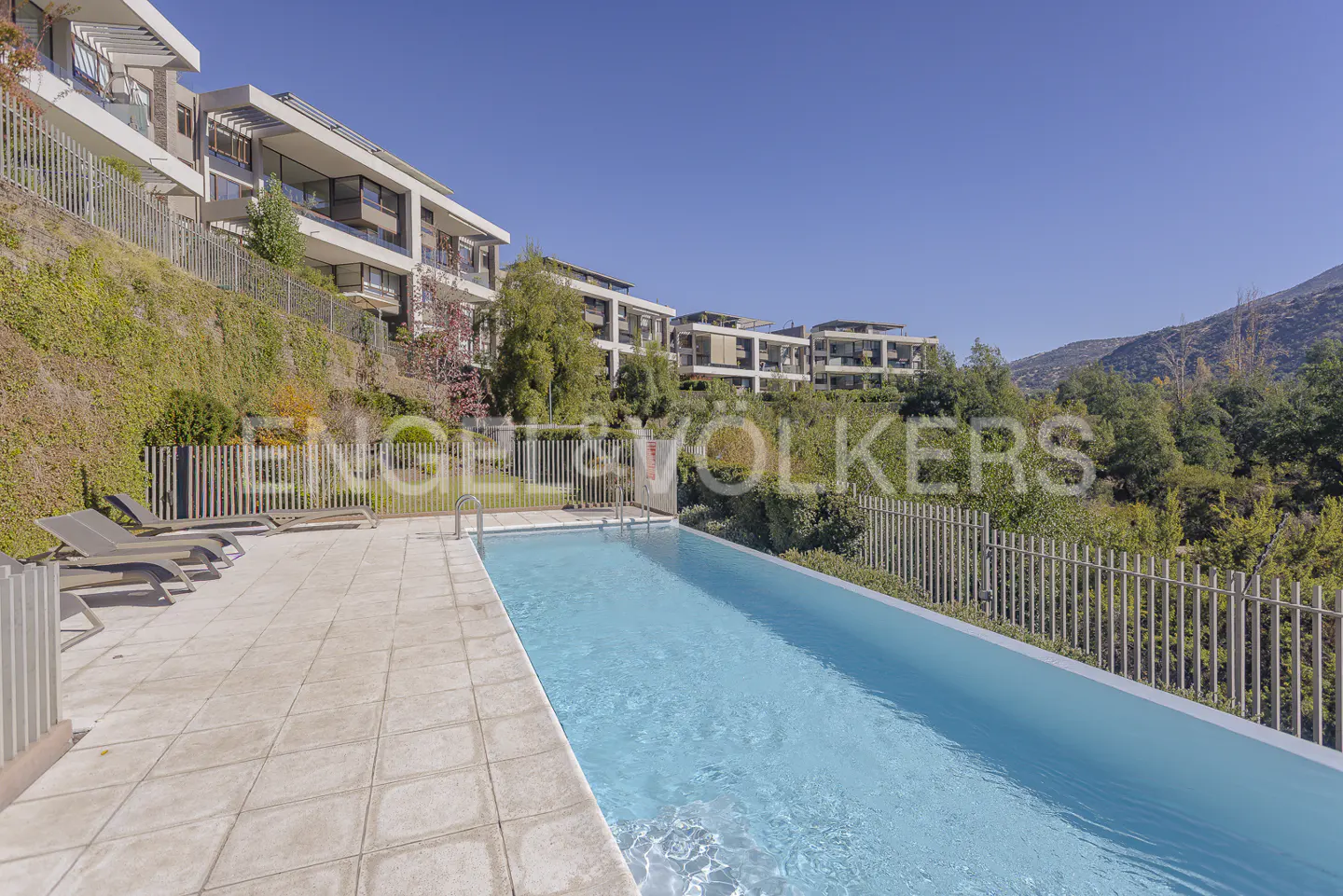 Outdoor pool with lounge chairs and modern hillside condos under a clear blue sky. Engel & Völkers logo overlay.