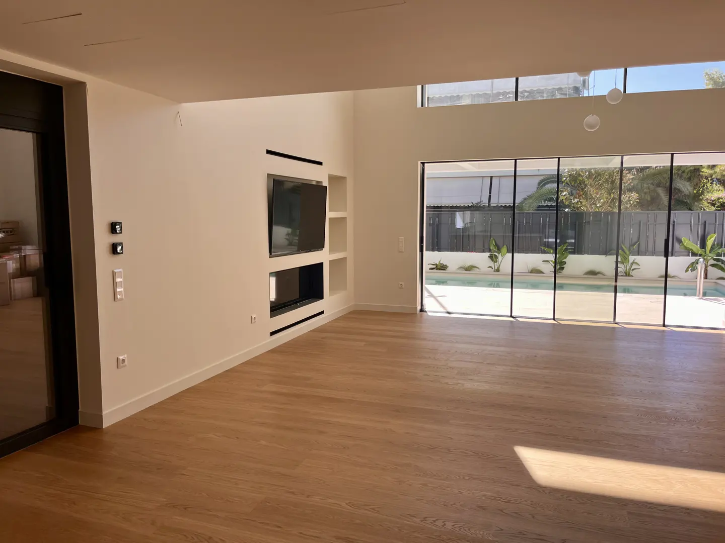Bright, modern living room with wood floors, white walls, and a large window overlooking a pool. A TV is mounted above a fireplace.