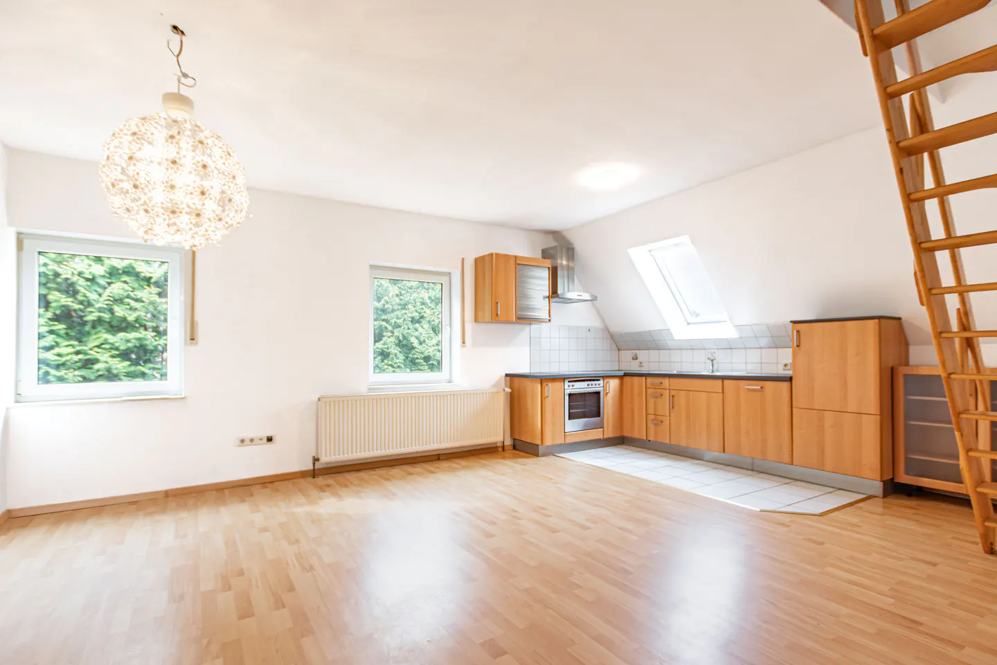 Bright, empty apartment with wood floors, white walls, and a kitchen area. A wooden ladder leads to an upper level. Two windows overlook green trees.