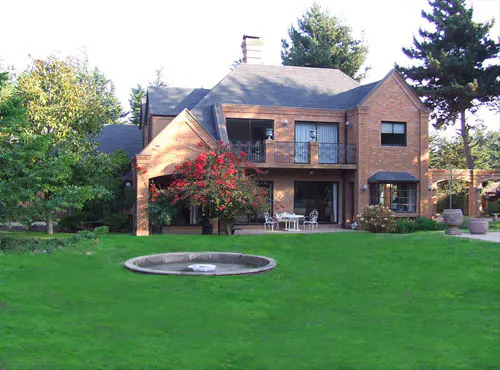 Two-story brick house with a dark roof, a balcony, and a large green lawn with a fountain.