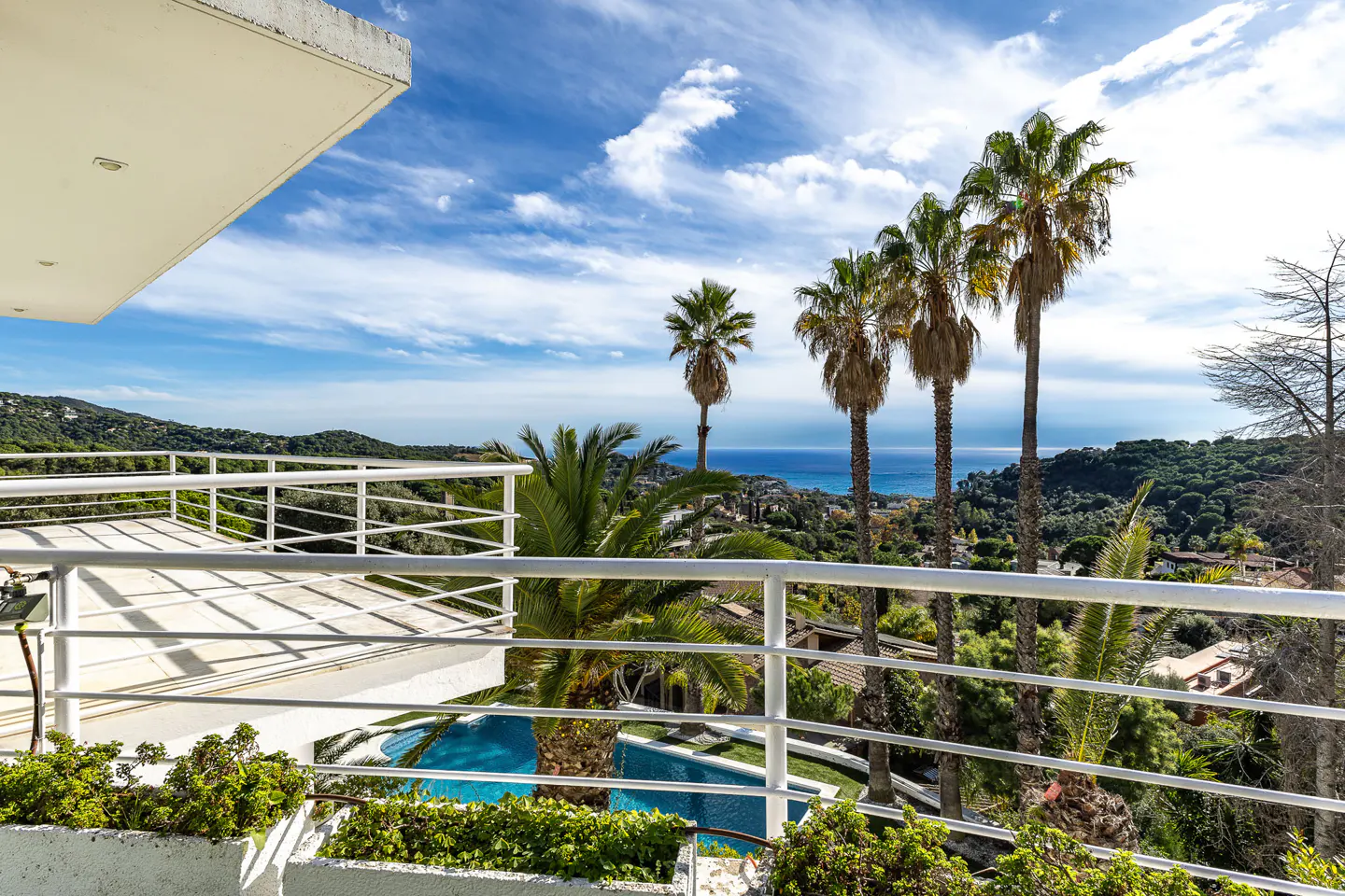 Balcony view of a pool, palm trees, and ocean. White railings frame the lush green landscape under a blue sky with scattered clouds.
