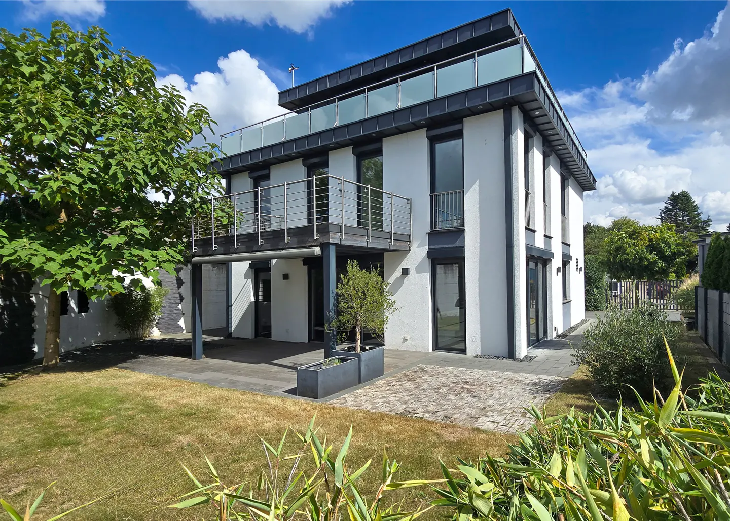 Modern two-story white house with black trim, balconies, and a rooftop terrace. Green lawn and trees surround the property.