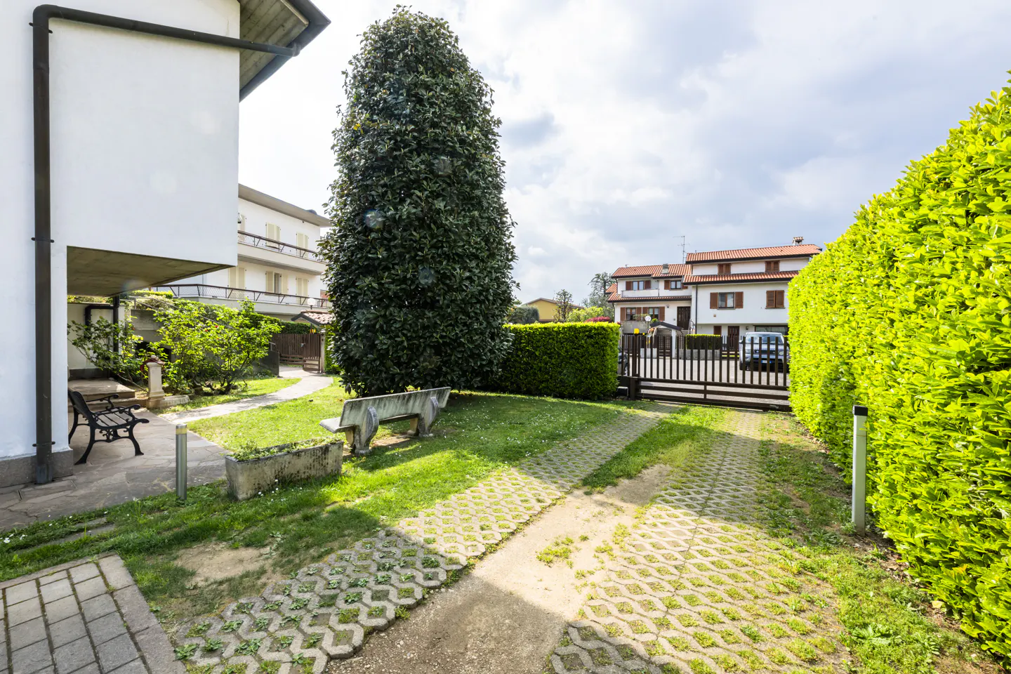 Exterior view of a home with a green lawn, tall tree, and a concrete bench. A hedge borders the property.