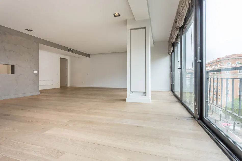 Bright, empty room with light wood floors, white walls, and large sliding glass doors to a balcony overlooking a city.