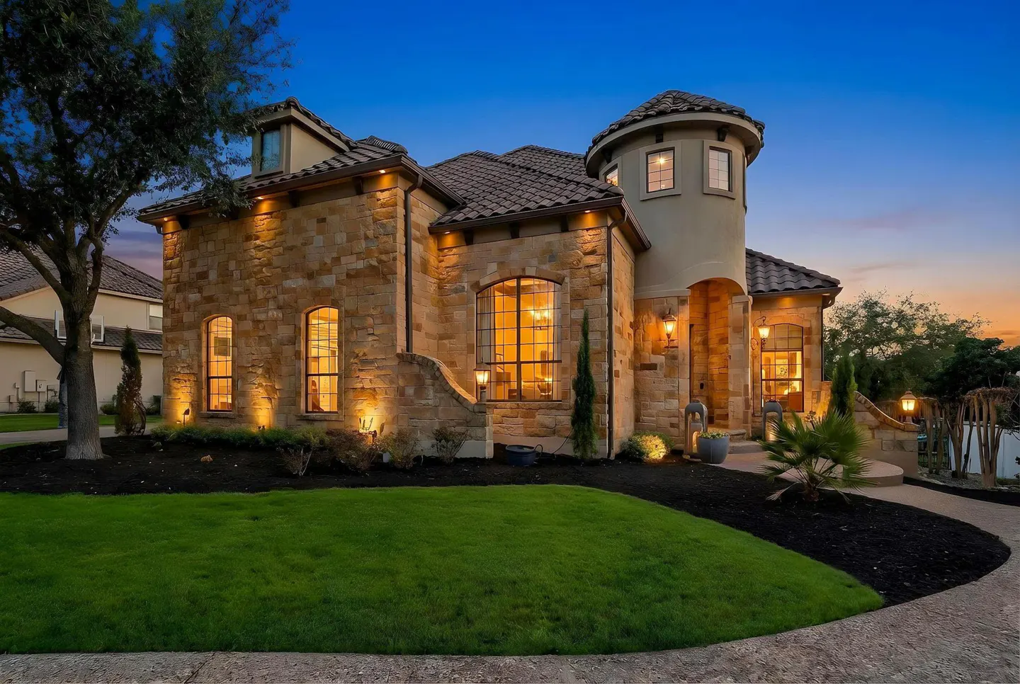 A two-story stone house with a turret, lit by warm lights at dusk, sits on a green lawn with dark mulch landscaping.