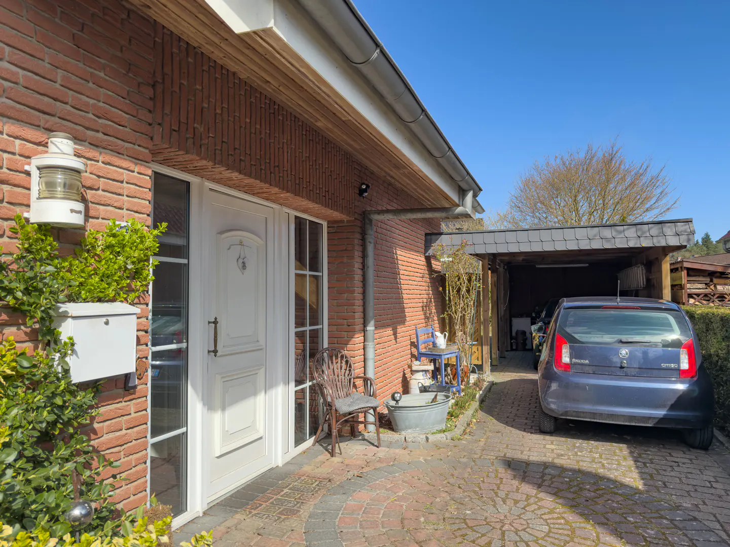 Exterior of a brick house with a white door, mailbox, and light fixture. A blue car is parked in the driveway under a carport.