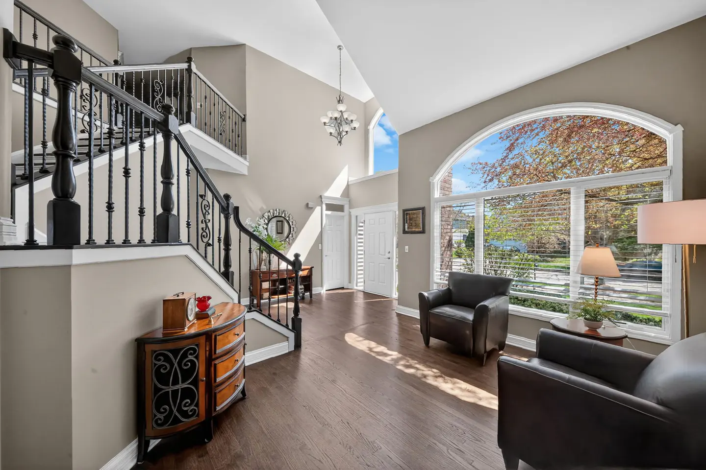 A bright foyer with dark wood floors, a staircase with black railings, and two leather armchairs near a large arched window.