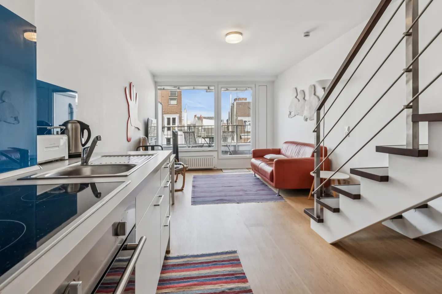 Bright apartment interior with white walls, wood floors, and a red leather sofa. A modern kitchen area is visible, along with stairs leading to an upper level.