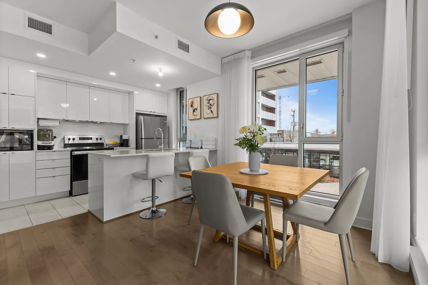 A modern condo interior with a light wood dining table, grey chairs, and a white kitchen with stainless steel appliances. A large window shows a city view.