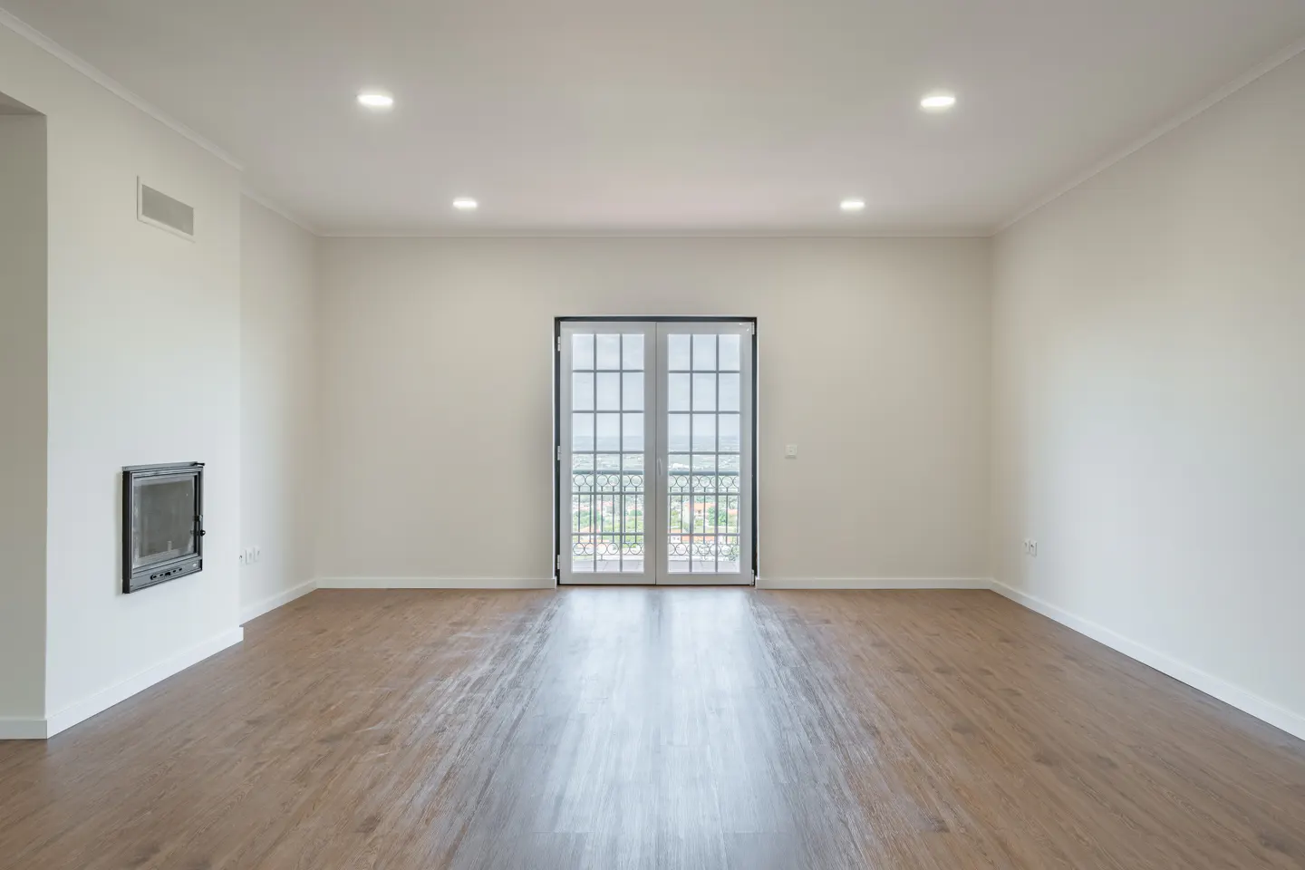 Bright, empty room with wood floors, white walls, and recessed lighting. French doors lead to a balcony with a view. A fireplace is on the left wall.