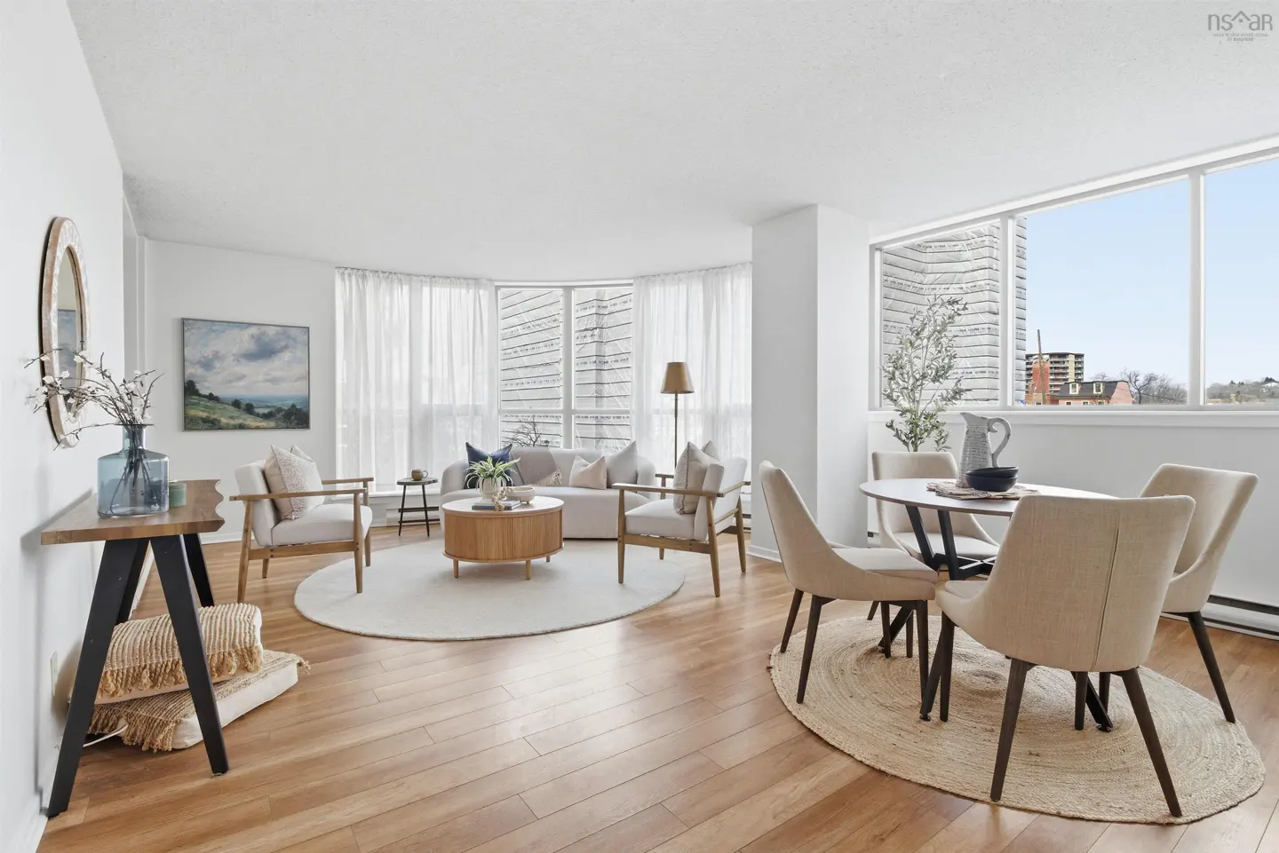 Bright, open-concept living and dining area with wood floors, white walls, and large windows. Beige sofa, chairs, and dining set.