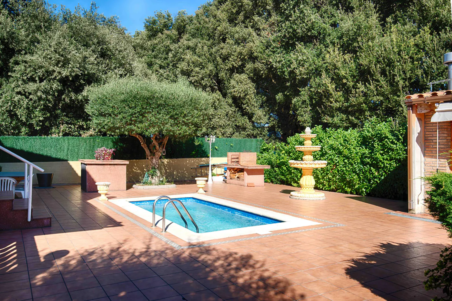 Backyard with a small blue pool, brown tile patio, yellow fountain, grill, and green trees.