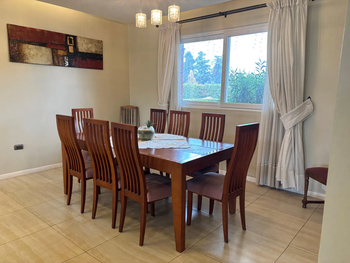 Dining room with a long wooden table and eight chairs, lit by a chandelier. A window with white curtains overlooks a green outdoor space.
