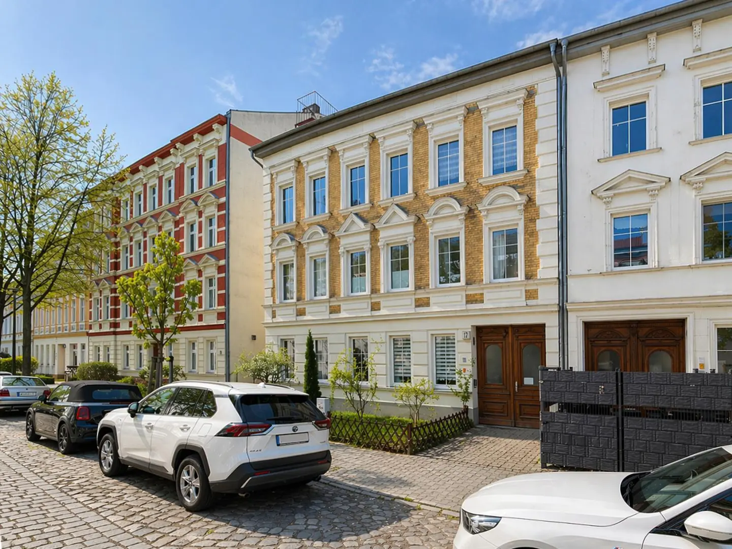 Street view of three ornate buildings with cars parked on a cobblestone street under a blue sky.