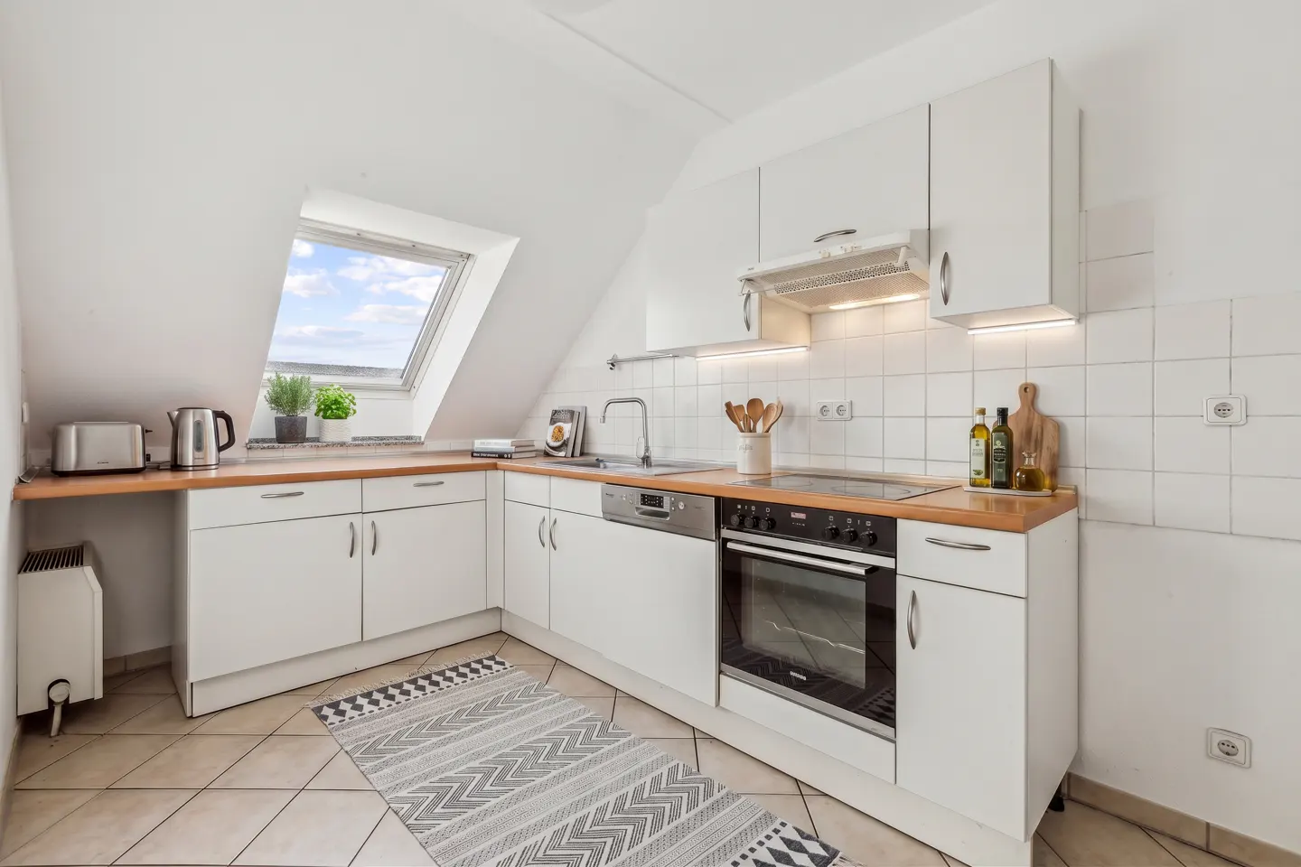 Bright kitchen with white cabinets, wood countertops, and a skylight. Appliances include an oven, dishwasher, and toaster. A patterned rug lies on the tiled floor.