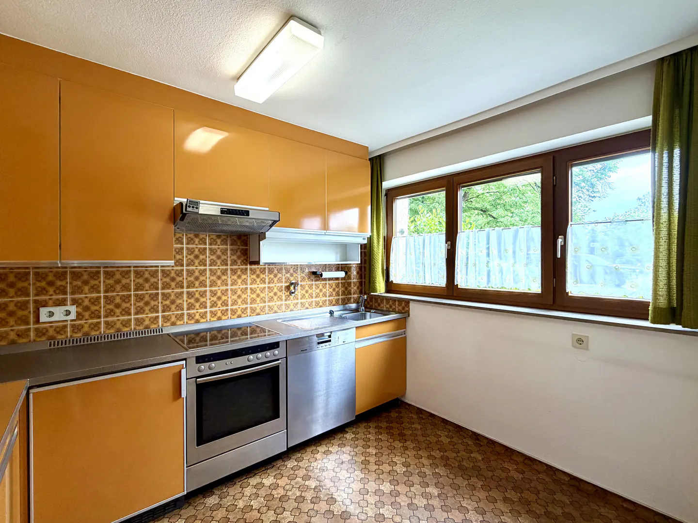 A retro kitchen with orange cabinets, stainless steel appliances, and a brown tiled backsplash. A window with green curtains overlooks a yard.