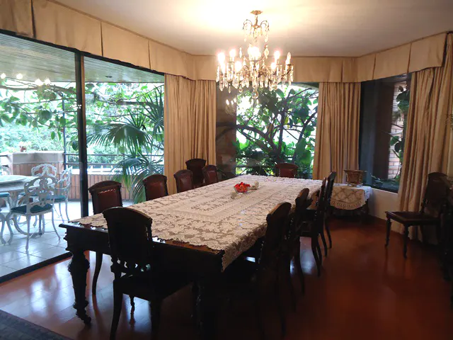 A formal dining room with a long table covered in a white lace tablecloth, surrounded by dark wood chairs. A crystal chandelier hangs above, and large windows offer views of lush greenery.