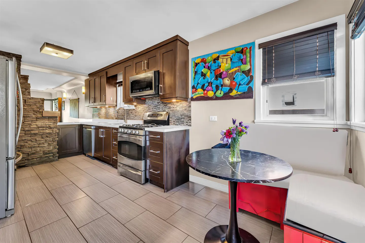 A kitchen with brown cabinets, stainless steel appliances, and a black marble table with flowers. A colorful painting hangs on the wall.